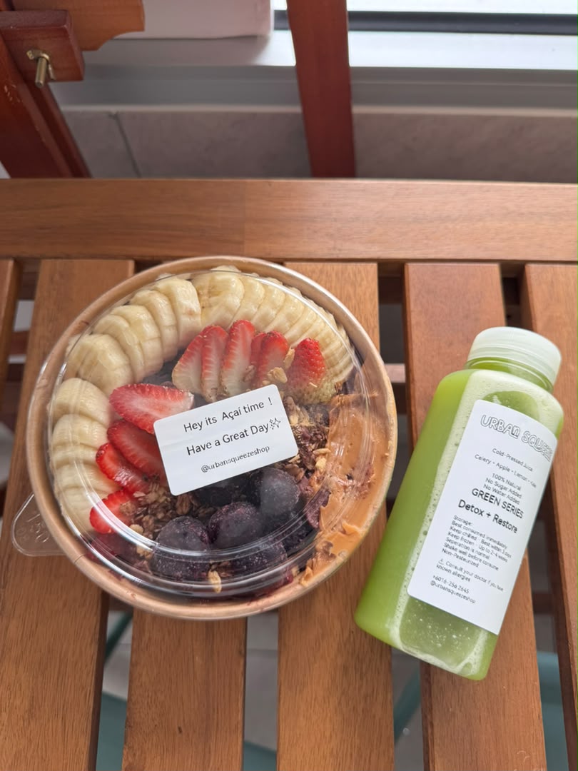 Açaí bowl topped with banana, strawberries and granola beside a Green Series juice on a wooden table