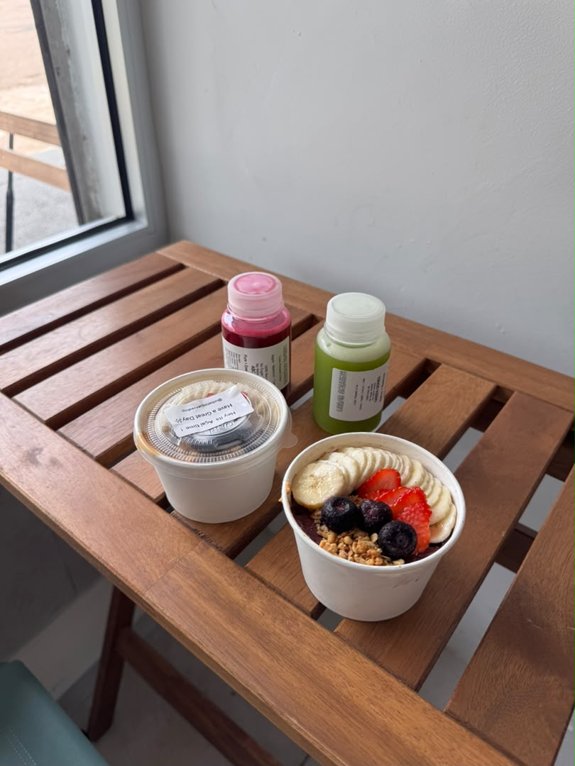 An open açaí bowl and a lidded bowl with red and green juice bottles on a wooden table